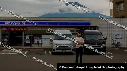 A young man stands with arms crossed in a convenience store parking lot with Mount Fuji rising dramatically in the background.
