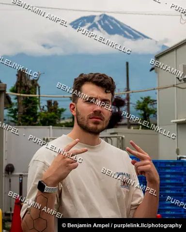 A young man makes peace signs with both hands while posing for a portrait with the snow capped peak of Mount Fuji visible behind him.
