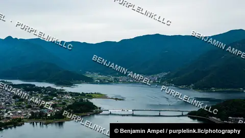 A high angle view shows the town of Kawaguchiko nestled beside a calm lake with a bridge crossing the water, surrounded by forested mountains.