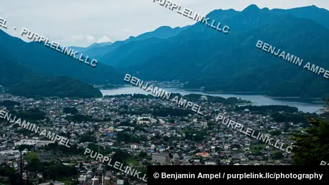 A hillside viewpoint overlooks the town of Kawaguchiko spread along the shore of a lake, surrounded by dense green mountains under an overcast sky.