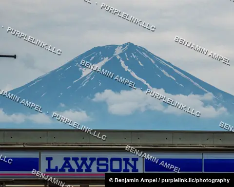 The snow streaked peak of Mount Fuji rises dramatically above the blue and white sign of a Lawson convenience store.