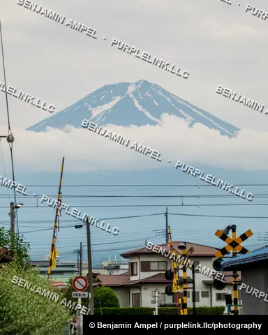 Mount Fuji's snow-streaked summit emerges above cloud cover, framed by rooftops, power lines, and a railway crossing in a small Japanese town.
