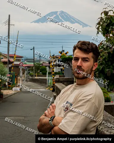 A man with a beard and tattoos stands with arms crossed on a stone wall, framed by a distant railway crossing and Mount Fuji rising above a Japanese town.