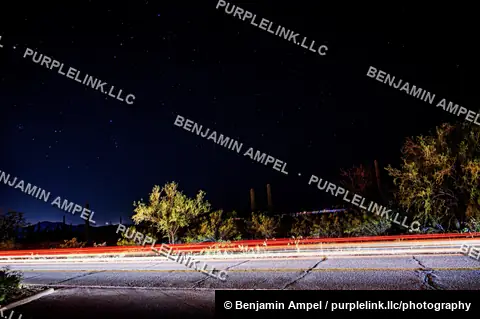 Long exposure captures a starry night sky and streaking vehicle light trails along a desert road in Arizona.