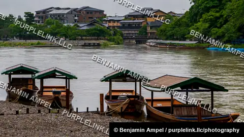 Traditional roofed wooden boats sit moored along the gravel bank of the Katsura River in Arashiyama, Kyoto, with riverside buildings across the water.