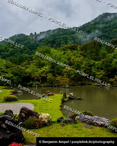 A tranquil Japanese garden pond bordered by moss-covered rocks and flowering shrubs sits beneath a mist-covered forested mountainside near Kyoto, Japan.