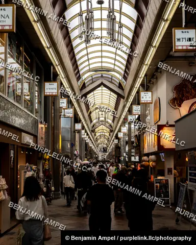 Shoppers walk through a long covered arcade lined with illuminated storefront signs and glass-roofed archways in Kyoto, Japan.