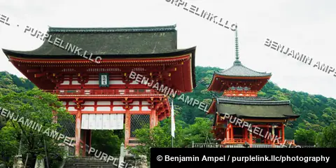 The vermillion Nio-mon gate and adjacent three-story pagoda of Kiyomizu-dera temple rise above lush green hillside forest in Kyoto, Japan.