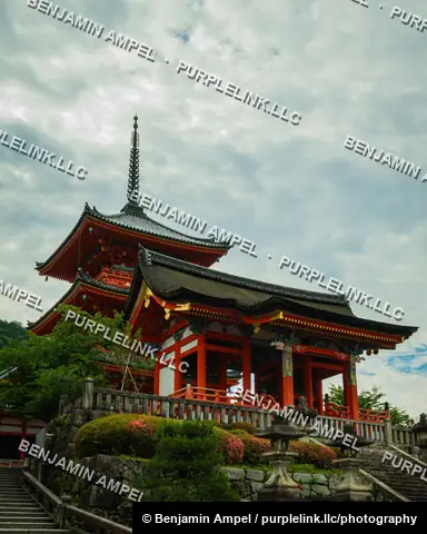 The layered three-story pagoda and main gate of Kiyomizu-dera temple stand atop stone terraces beneath a dramatic cloudy sky in Kyoto, Japan.