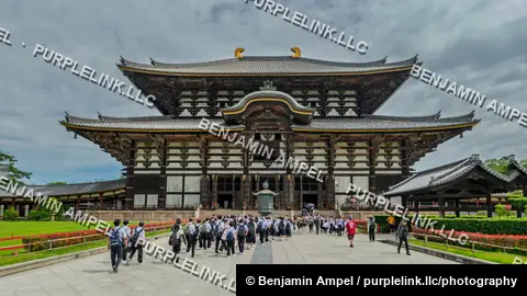 Groups of visitors and schoolchildren walk toward the massive Great Buddha Hall of Todai-ji temple beneath a cloudy sky in Nara, Japan.
