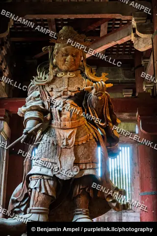 A weathered wooden guardian statue in ornate armor stands inside the wooden hall of Todai-ji temple in Nara, Japan.