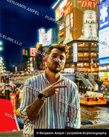A young man gestures a peace sign while standing before the glowing Asahi and Glico billboards of Dotonbori at night in Osaka, Japan.