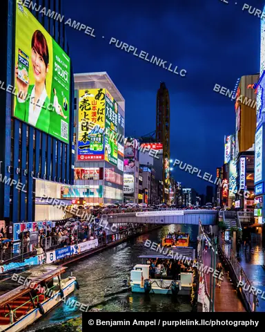 Tour boats glide along the Dotonbori canal at dusk beneath glowing storefronts, billboards, and a crowded pedestrian bridge in Osaka, Japan.
