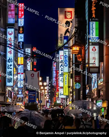 Pedestrians carry umbrellas through a narrow street lined with tall illuminated signs, including a FamilyMart and Starbucks logo, in Osaka's Dotonbori district.