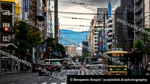A streetcar rolls through a busy Hiroshima street lined with illuminated shop signs and traffic beneath overcast skies.