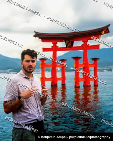 Miyajima, Japan - June 2025: A young man makes a peace sign while standing in front of the iconic red torii gate rising from the sea.