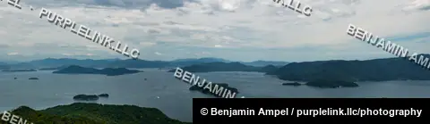 A wide panorama shows forested islands scattered across the calm waters of the Seto Inland Sea under a cloudy sky.