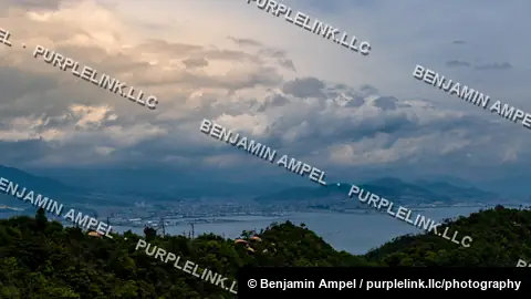 Storm clouds glow with evening light over Hiroshima's coastal city and bay, seen from a forested mountain ridge.