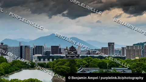 Dark storm clouds loom over Hiroshima Castle and the surrounding city skyline, with mountains in the distance.