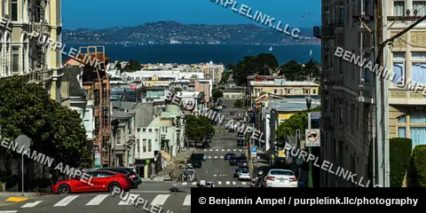 A steep San Francisco street lined with cars and buildings descends toward the bay, with sailboats and hills visible in the distance.