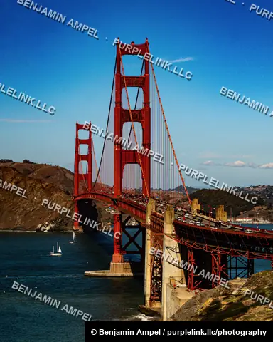 The Golden Gate Bridge spans the entrance to San Francisco Bay, with sailboats crossing the water below and coastal hills in the background.