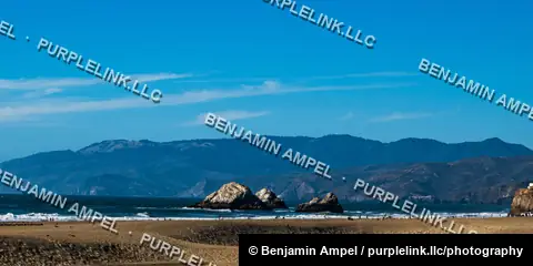 Rocky sea stacks rise from the surf off Ocean Beach with distant coastal mountains under a clear blue sky.