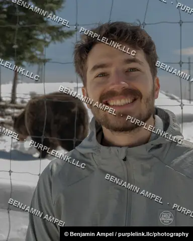 Vancouver, Canada - April 2026: A visitor poses for a photo with a grizzly bear grazing behind a wire fence at the Grouse Mountain wildlife refuge.