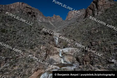 A tiered waterfall drops through a rocky Sonoran Desert canyon lined with saguaro cacti beneath a deep blue sky.