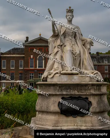London, England - October 2025: A stone statue of a seated Queen Victoria holding a scepter stands before the red-brick facade of Kensington Palace.