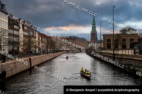 A rowing crew paddles along a historic city canal lined with colorful townhouses under a dramatic cloudy sky at dusk.