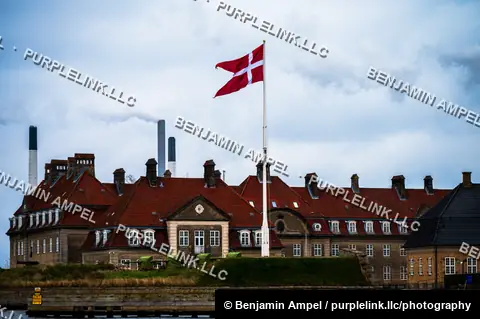 A row of historic red-roofed brick barracks buildings flies a large Danish flag, with industrial smokestacks rising in the background.