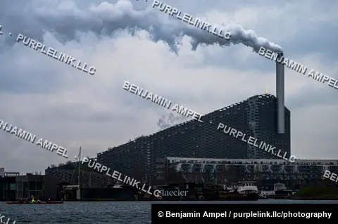 Copenhagen, Denmark - December 2025: Steam rises from the smokestack of the Amager Bakke waste-to-energy plant, seen across the harbor with rowers and houseboats in the foreground.