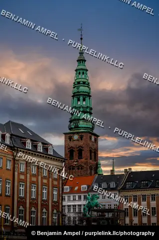 Copenhagen, Denmark - December 2025: The green spire of Nikolaj Church rises above an equestrian statue and historic rooftops as sunset colors the clouds.