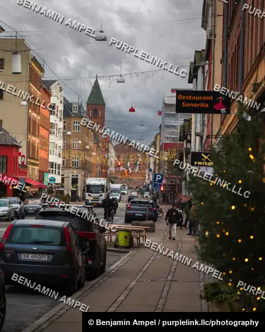 Copenhagen, Denmark - December 2025: Cars line a rainy city street strung with Christmas lights, with a church spire visible past neon restaurant signs and parked cars.