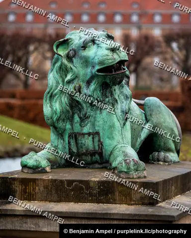 A weathered green bronze lion statue sits on a stone pedestal, with a red-roofed historic building softly blurred in the background.