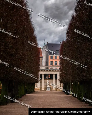 Copenhagen, Denmark - December 2025: A symmetrical hedge-lined path leads toward a classical colonnade and statue framed by a pink townhouse under a moody sky.