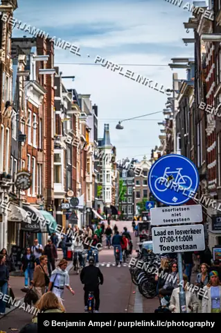 Amsterdam, Netherlands - June 2026: Pedestrians and cyclists fill a narrow shopping street beneath a bicycle route sign.