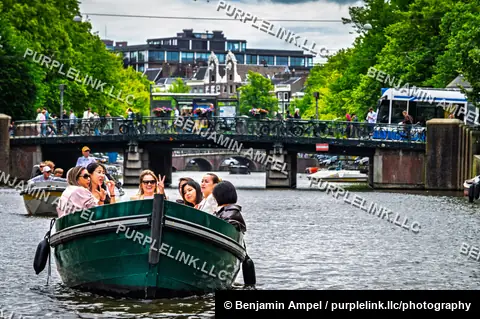 Amsterdam, Netherlands - June 2026: Passengers pose and wave from a canal boat passing beneath a bridge lined with cyclists.