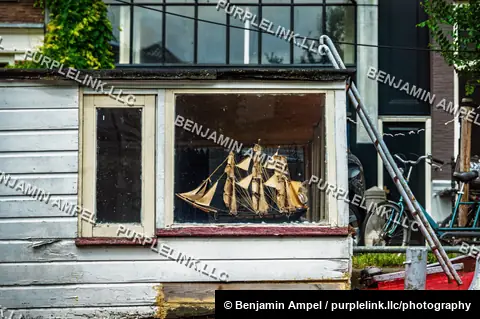 A detailed model sailing ship sits in the wooden window box of a weathered Amsterdam houseboat.