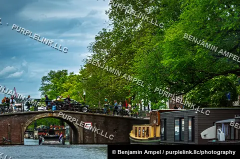 Amsterdam, Netherlands - June 2026: Houseboats line a tree-shaded canal beneath a stone bridge crossed by pedestrians and cyclists.