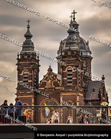 Amsterdam, Netherlands - June 2026: Travelers with luggage cross a bridge in front of the twin spires of the Basilica of Saint Nicholas.