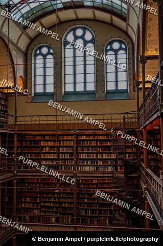 Tiered wooden bookshelves and a spiral staircase rise beneath tall arched windows in the Rijksmuseum's historic library.