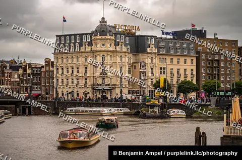 Amsterdam, Netherlands - June 2026: Tour boats cruise past the historic Hotel Victoria near Amsterdam Centraal on an overcast day.