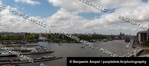 A wide panorama shows moored houseboats along the Amstel river with the Amsterdam skyline and church spires in the distance.
