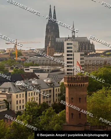 Cologne Cathedral's Gothic spires and a brick medieval tower rise above the city's rooftops at dusk.
