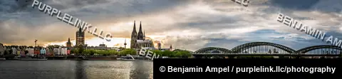 A wide panorama of Cologne's cathedral, old town, and the Hohenzollern railway bridge spanning the Rhine river at sunset.
