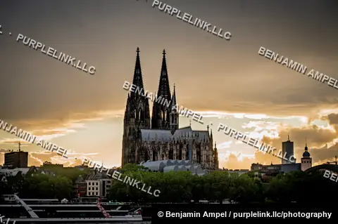 The twin Gothic spires of Cologne Cathedral rise in silhouette above the Rhine as a passenger ship passes at sunset.