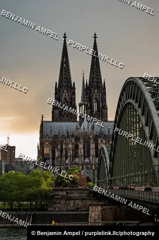 The Gothic towers of Cologne Cathedral rise beyond the Hohenzollern railway bridge as pedestrians cross over the Rhine at dusk.