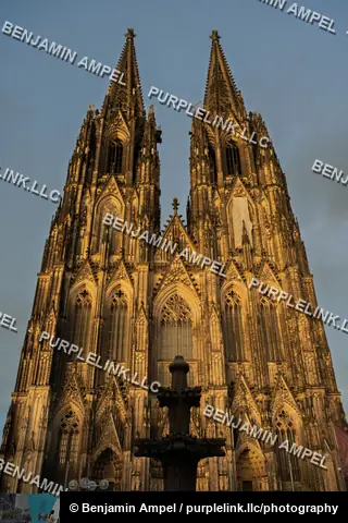 The ornate Gothic facade and twin spires of Cologne Cathedral glow in warm evening light against a dramatic sky.