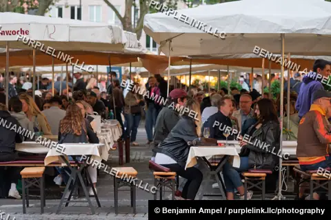 Cologne, Germany - June 2026: Visitors gather at wine glasses and tables under white market tents during an evening street event.